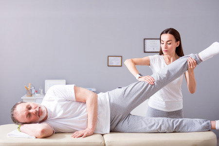 Personal treatment. Glad male patient lying down with female concentrated and serious physiotherapist performing some stretch exercises on mans legの写真素材