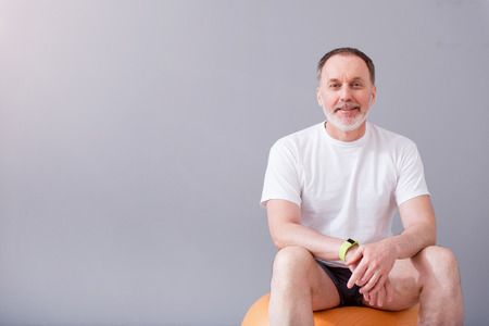 Sporty man. Smiling and cheerful man sitting on a gym ball during physiotherapy and being on a isolated grey backgroundの写真素材