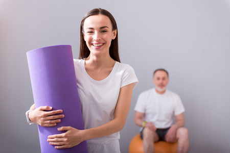 Smiling therapist. Cheerful female physiotherapist holding a mat for exercises and a male patient ...