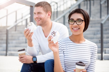 So tasty. Content and delighted young man and young woman being outdoors sitting on the stairs while eating sweets and drinking coffeeの写真素材
