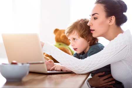 Thats interesting. Young pretty mother and her charming little son looking attentively at the laptop while sitting at the tableの写真素材