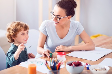 I like painting. Fascinated little boy holding a brush and looking in front of him while his mother holding a jar of painting and looking at himの写真素材