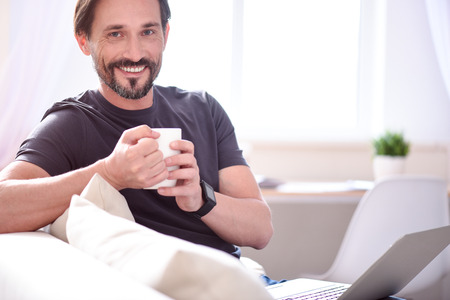 At last I am at home. Smiling mature bearded man looking at the camera while sitting and holding a...