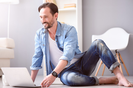 Feel yourself comfortable. Delighted mature man smiling while sitting on the floor and using his laptopの写真素材