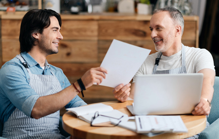 Always together. Merry and smiling young man and his cheerful father using laptop, holding paper together while being at homeの写真素材