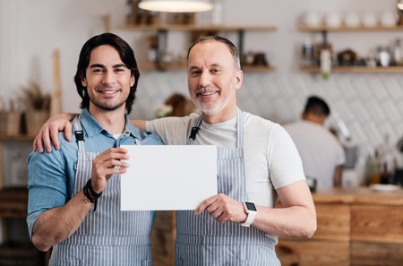 Start your business. Positive and happy male cafe workers in apron holding a sheet of white paper while arming each other and smiling at a camera, standing in a cafeの写真素材