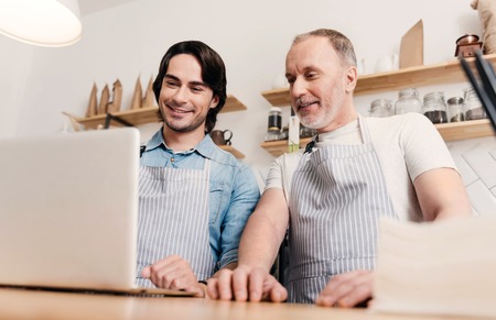 Technologies at work. Cheerful and positive two male cafe workers using laptop at workの写真素材