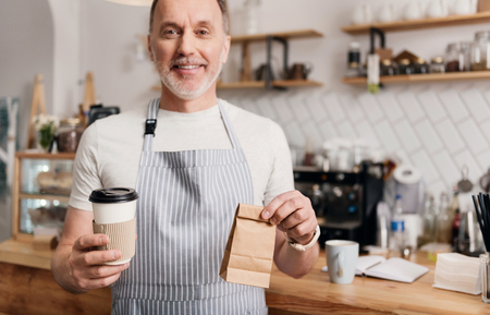 You are welcome. Smiling and cheerful male cafe worker in apron holding coffee to go and little lunch bagの写真素材