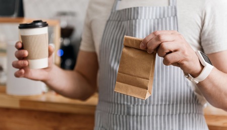 Take it please. Cropped image of hands of male cafe worker in apron holding coffee to go and little...