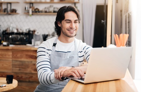 Ways of communication. Happy and positive male cafe worker sitting at the bar and using laptop while the break at workの写真素材