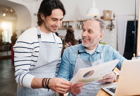 Our results. Smiling and content male senior cafe worker sitting at the bar, using laptop while showing some papers for his younger coworkerの写真素材