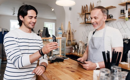 Nice communication. Smiling and cheerful client showing something on his mobile phone for content and merry male barista with digital tabletの写真素材