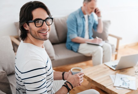 Freelancers team. Smiling young man holding a cup of coffee and looking at a camera with another man sitting, talking per phone and noting something in a backgroundの写真素材