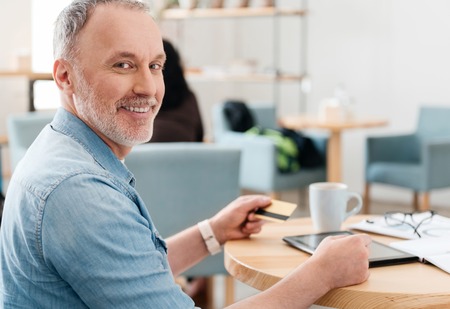 My usual day. Smiling and positive man using digital tablet and credit card while drinking coffee and looking at a cameraの写真素材