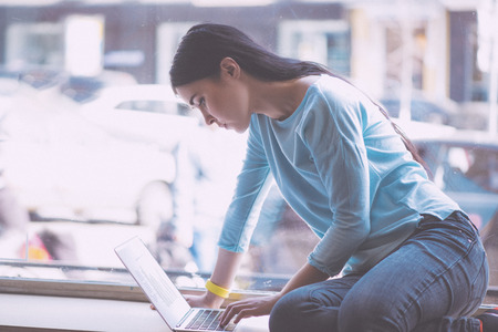 Involved in work. Concentrated and confident young woman sitting on a window sill and using laptop while being in a cafeの写真素材