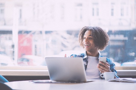 Wow. Happy and cheerful young man using mobile phone and laptop while being in a cafeの写真素材