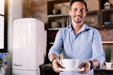 Take it. Handsome bearded man smiling and giving you a cup of delicious coffee while holding it with both handsの写真素材