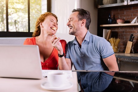 Eat it. Ecstatic man laughing at his wife eating a croissant while sitting at the table in the kitchenの写真素材