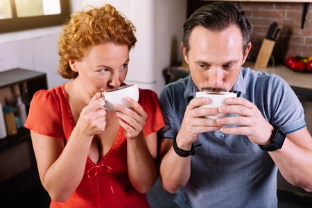 Funny morning. Cute middle aged couple drinking coffee and holding cups near mouthの写真素材