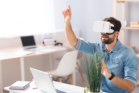 Hey. Pleasant emotional man holding his hand up and using virtual reality glasses while sitting at...