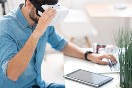 Ready to work. Pleasant man wearing virtual reality glasses and using tablet while sitting at the...