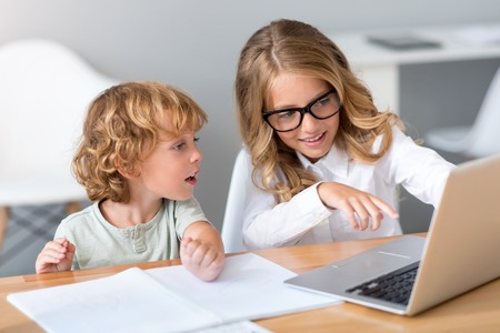 Have a look. Beautiful little girl with glasses showing something to the interested little child while pointing at the screen of the laptopの写真素材