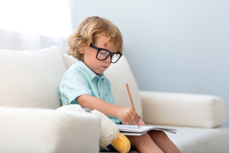 I have a dream. Little concentrated boy wearing glasses and drawing while sitting on the couchの写真素材