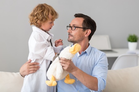 Here is a new patient. Serious man giving to his little cute doctor in a hospital gown a toy while looking at himの写真素材