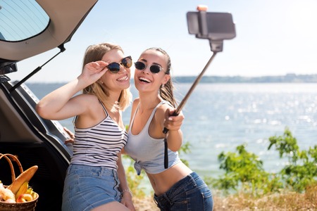 Life fully. Overjoyed young smiling friends making selfies and standing near car while resting near...