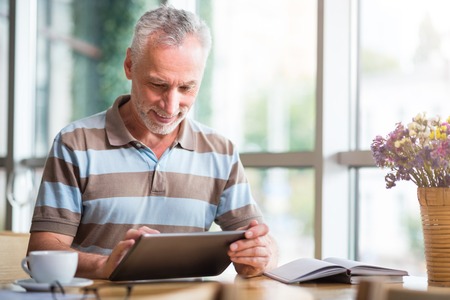 Modern user, Pleasant positive senior man sitting at the table and smiling while using tablet
