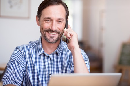 Always connected. Delighted and content man using laptop and talking at phone sitting at the table