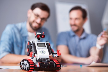 Nice device. Selective focus of robot standing on the table while cheerful colleagues sitting in the...