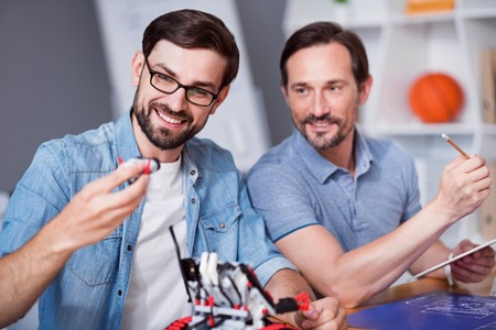Find right detail. Positive content colleagues sitting at the table and using robot while working on...