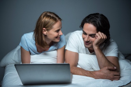 Rest together. Cheerful young loving couple lying in bed and expressing joy while using laptopの写真素材
