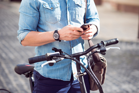 Everyday life. Close up of cell phone in hands of pleasant man holding a cellphone and standing with the bicycle in the streetの写真素材