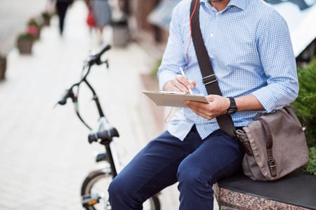 Put it down. Pleasant man sitting in the street near his bicycle and holding folder while making notesの写真素材