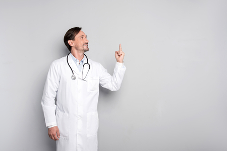 I found it. Handsome young inspired doctor pointing his finger up while thinking and wearing his stethoscope by the grey background.の写真素材