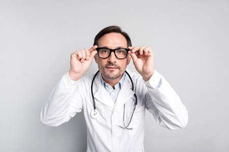 Oh my God. Handsome young doctor staring and touching his glasses while standing against grey...