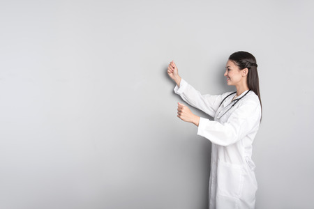 Do it this way. Cheerful professional female doctor holding hands aside and smiling while standing isolated on grey backgroundの写真素材