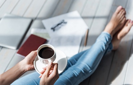 Small pause. Top view of lady sitting barefoot on grey wooden floor and holding full coffee cup.の写真素材