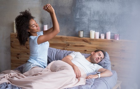 Time to wake up. Sleepy beautiful girl stretching herself after waking up while her lovely husband is sleeping.の写真素材