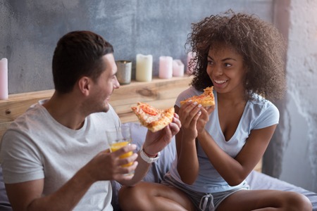 Snack time. Young delighted sweet couple eating pizza and drinking juice while sitting in bed together happily.の写真素材