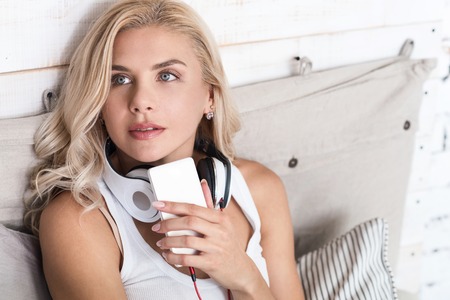 Deep thoughts. Pretty blond lady wearing headphones, holding her new smartphone and looking aside.の写真素材