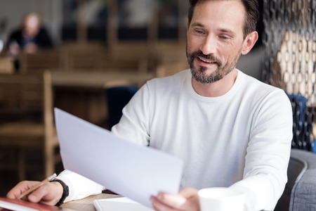 Attentively reading. Handsome bearded man sitting in office reading important document and happily smiling.の写真素材
