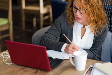 Serious information. Beautiful ginger-haired woman sitting in cafe and taking notes while looking at modern laptop.の写真素材