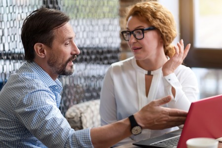 Discussing new project. Middle-aged bearded man pointing at laptop screen while discussing business deals with talking ginger woman.の写真素材