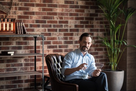 Enjoying his job. Happy delighted handsome man sitting in the armchair and smiling while working on the tabletの写真素材