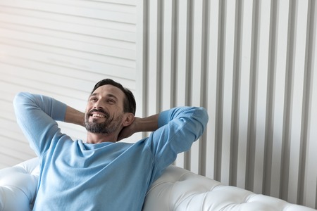 Feeling great. Nice cheerful bearded man holding his hands behind the head and smiling while being in a wonderful moodの写真素材