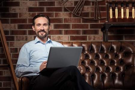 Expressing emotions. Happy positive stylish man smiling and looking in front of him while sitting on the sofaの写真素材