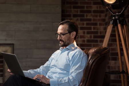Care about your eyesight. Confident optimistic nice businessman sitting at the laptop and looking at the screen while wearing glassesの写真素材